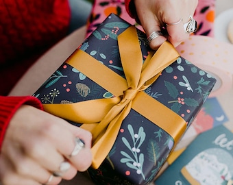May include: A wrapped gift with navy blue wrapping paper featuring a botanical pattern of green leaves, red berries, and pinecones. The gift is tied with a wide, golden-yellow ribbon. Other gifts and tags are in the background.