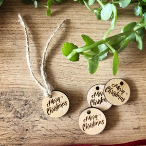 Puede incluir: Cuatro etiquetas circulares de madera clara, cada una con el texto Merry Christmas grabado con láser en letra cursiva. Una etiqueta tiene un trozo de cuerda de yute natural enhebrado a través de un pequeño orificio, mientras las otras tres están sueltas.