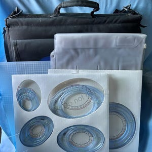 May include: A black carrying case with a handle, a clear plastic envelope, and several blue oval-shaped templates in white packaging. The templates have red and blue markings. A blue cutting mat is also visible.