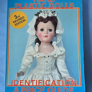 Peut inclure: Un livre intitulé "Hard Plastic Dolls" avec une poupée en robe de mariée sur la couverture. La couverture bleue porte le texte "Identification & Price Guide" et "3rd Revised Edition". La poupée porte un voile blanc et une coiffe florale.