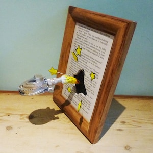 Könnte beinhalten: Ein silbernes und weißes Modell eines Raumschiffs bricht durch ein Loch in einem Buch und hinterlässt eine Spur gelber Flammen. Das Buch befindet sich in einem Holzrahmen und die Seite ist mit Text gefüllt. Der Text lautet: "Er kam mir näher, fuhr er fort. "Und vielleicht hat er nur die Jacke..."