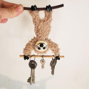 Puede incluir: Un llavero de perezoso de peluche marrón claro, colgado boca abajo de una rama de madera. El perezoso tiene un ojo de botón y sostiene una clavija de madera con tres llaves adjuntas. El llavero es un artículo decorativo.