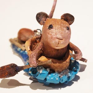 May include: A small, brown mouse figurine is sitting in a walnut shell boat. The boat is surrounded by blue and white beads, and the mouse is holding a small wooden paddle.