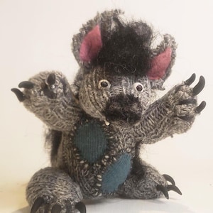 May include: A handmade knitted monster doll with gray and black yarn. The monster has pink ears, black claws, and a teal patch on its chest. The doll is posed with its arms outstretched.