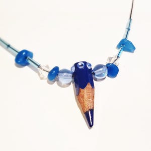 May include: A handcrafted necklace with a pencil pendant, dark blue at the top and natural wood at the base. The necklace includes blue and clear beads, strung on a silver wire. The pencil pendant is approximately 2.5 cm long.