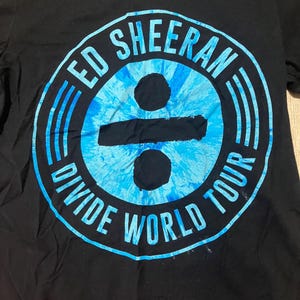 Könnte beinhalten: Schwarzes T-Shirt mit blau-weißem Batik-Design. Das Design zeigt den Text "ED SHEERAN DIVIDE WORLD TOUR" in kreisförmiger Anordnung. Das Shirt zeigt den Namen des Künstlers und Tourdetails.