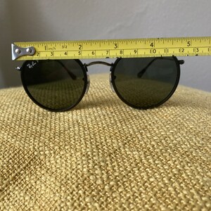 Op de afbeelding: Zwarte ronde Ray-Ban zonnebril met donkere glazen. De zonnebril wordt gemeten met een meetlint en is ongeveer 10 cm breed.