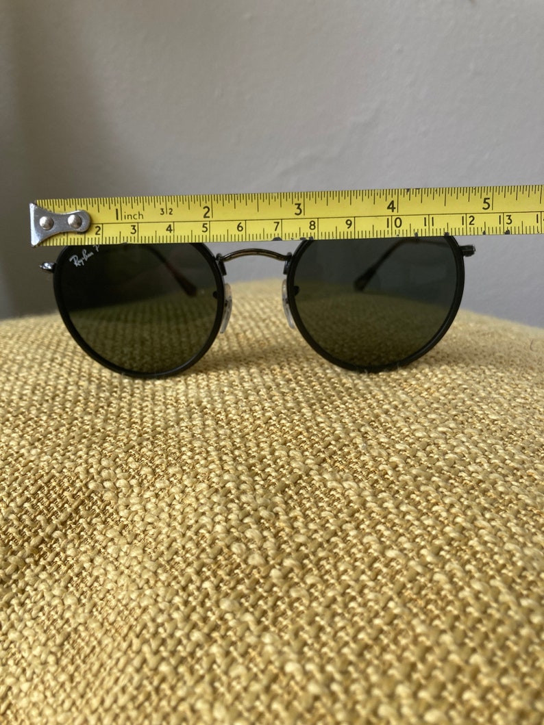 Op de afbeelding: Zwarte ronde Ray-Ban zonnebril met donkere glazen. De zonnebril wordt gemeten met een meetlint en is ongeveer 10 cm breed.
