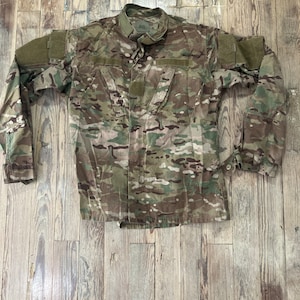 Op de afbeelding: Een lang mouw camouflage shirt in groen, bruin en tan met een knoopsluiting en klittenband patches op de mouwen.