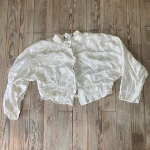 May include: A white, crinkled, cropped blouse with a button-up front and long sleeves.