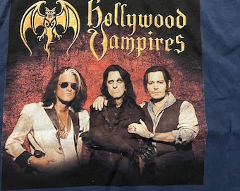 May include: A blue t-shirt featuring a graphic of the band "Hollywood Vampires". The graphic depicts a bat with a skull, and three men in front of a red background. The band's name is written in gold lettering.
