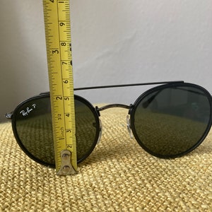 Op de afbeelding: Zwarte ronde Ray-Ban zonnebril met groene glazen. De zonnebril wordt gemeten met een gele liniaal, wat aangeeft dat de breedte van de glazen ongeveer 6,5 cm is.