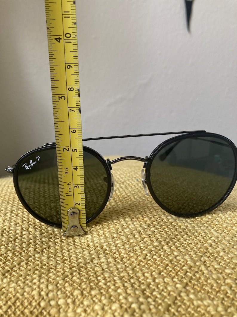 Op de afbeelding: Zwarte ronde Ray-Ban zonnebril met groene glazen. De zonnebril wordt gemeten met een gele liniaal, wat aangeeft dat de breedte van de glazen ongeveer 6,5 cm is.
