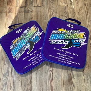 Può includere: Due cuscini per sedili viola e blu con il testo "Devil Rays Inaugural Season 1998" e il logo GTE. I cuscini presentano una mazza da baseball e un cappello da strega.
