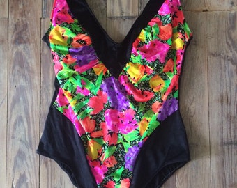 Puede incluir: Un bañador negro de una pieza con estampado floral en tonos rosa, amarillo, verde y naranja. El bañador tiene un escote en V y un ribete negro.