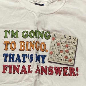 Op de afbeelding: Wit T-shirt met een kleurrijke afbeelding met de tekst "Ik ga naar bingo. Dat is mijn definitieve antwoord!" en een bingokaart met nummers.