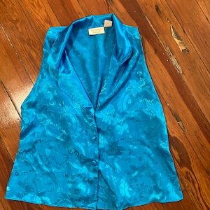 Puede incluir: Un top azul de satén sin mangas con un estampado floral y cierre de botones. El top tiene un cuello y la etiqueta "Victoria's Secret" es visible.