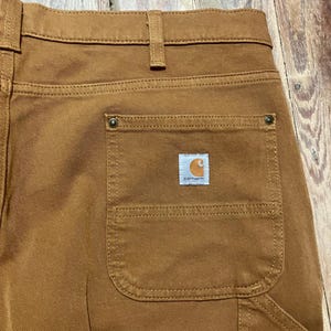 Puede incluir: Primer plano de pantalones de trabajo marrones con un bolsillo trasero y un pequeño parche con el logotipo de Carhartt. Los pantalones tienen un diseño clásico con costuras reforzadas y tejido duradero.