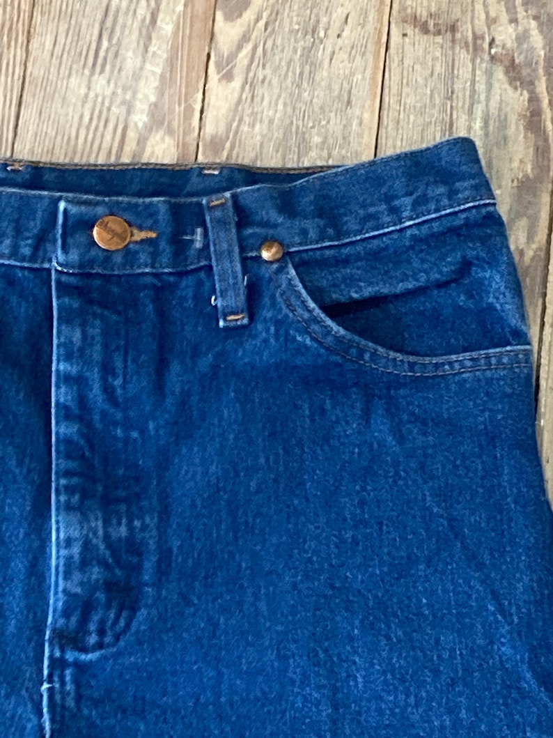 May include: Close-up of the top of a pair of blue denim jeans with a copper button and rivets. The jeans have a classic five-pocket style.