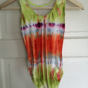 May include: A tie-dye leotard in shades of green, orange, purple, and blue hangs on a wooden hanger. The sleeveless garment features a scoop neck and a neon green strap across the back. The fabric is a vibrant mix of colors.