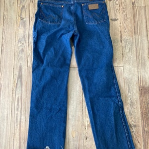 May include: A pair of blue denim jeans with a frayed hem. The jeans have a brown leather patch on the back with the Wrangler logo.