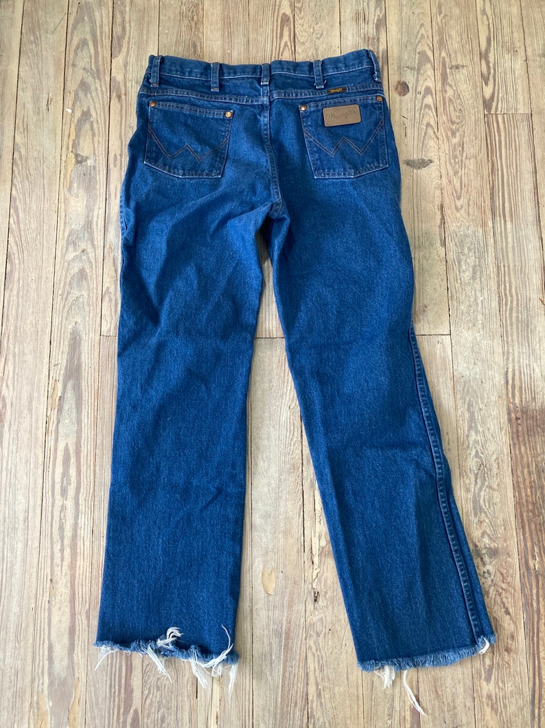 May include: A pair of blue denim jeans with a frayed hem. The jeans have a brown leather patch on the back with the Wrangler logo.