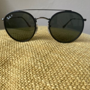 Op de afbeelding: Zwarte ronde Ray-Ban zonnebril met een dun metalen frame en donkere glazen.