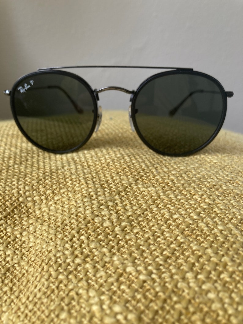 Op de afbeelding: Zwarte ronde Ray-Ban zonnebril met een dun metalen frame en donkere glazen.