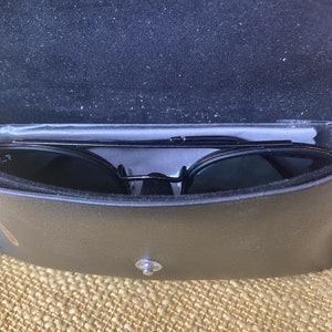 Op de afbeelding: Zwarte zonnebril in een zwarte leren etui. De etui heeft een zilveren knoopsluiting.