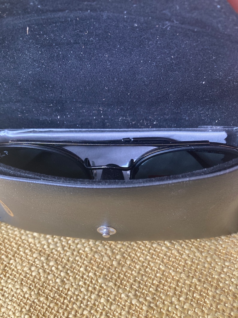 Op de afbeelding: Zwarte zonnebril in een zwarte leren etui. De etui heeft een zilveren knoopsluiting.