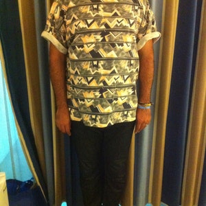 May include: A man wearing a short-sleeved shirt with a geometric pattern in shades of grey, white, and beige. He is wearing dark blue jeans and white trainers.