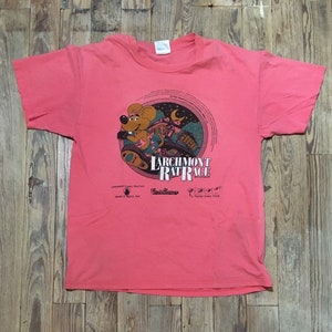 May include: A pink t-shirt with a graphic of a cartoon rat driving a car. The graphic is in a circle with the text "Larchmont Rat Race" inside. The shirt is made of cotton and has a crew neck.