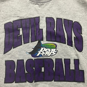 Pode incluir: Uma camiseta cinza com um gráfico roxo do texto "Devil Rays Baseball" com um logotipo roxo e azul de um peixe raia diabo com o texto "Tampa Bay Devil Rays" em um círculo.