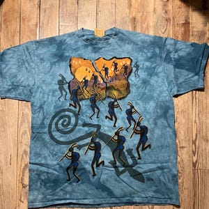 May include: Blue tie-dye t-shirt featuring a Kokopelli design. The graphic depicts silhouetted figures playing flutes, with a sun and spiral elements. The shirt is made of a soft material and has a crew neck.