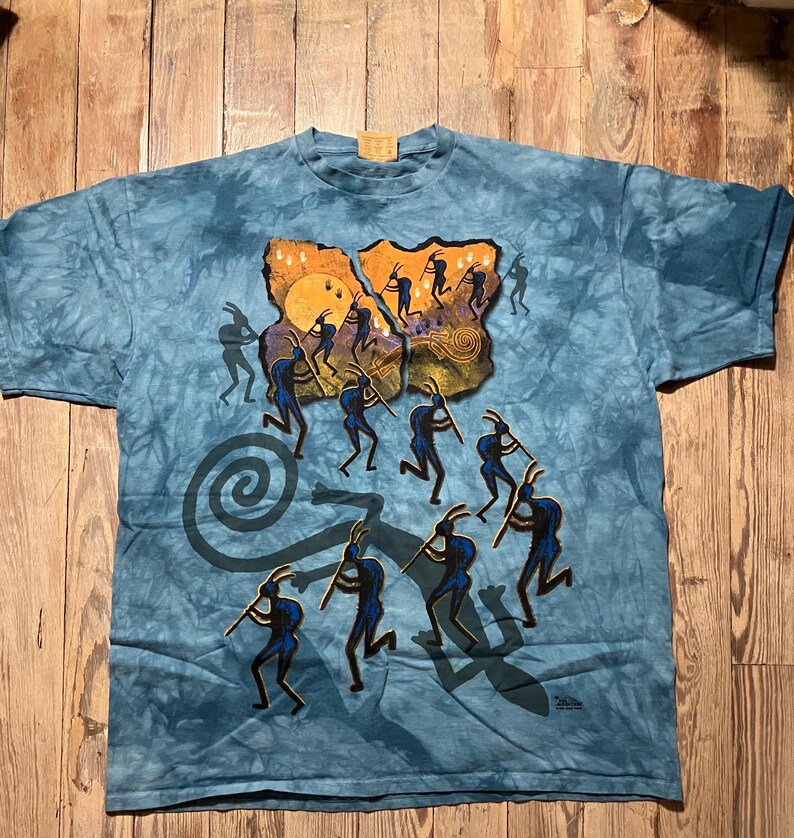 May include: Blue tie-dye t-shirt featuring a Kokopelli design. The graphic depicts silhouetted figures playing flutes, with a sun and spiral elements. The shirt is made of a soft material and has a crew neck.