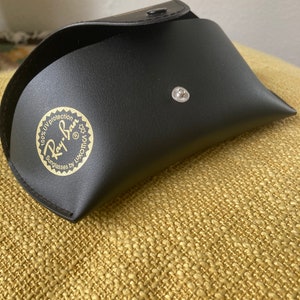 Op de afbeelding: Zwarte leren etui voor zonnebrillen met een gouden Ray-Ban logo met de tekst "100% UV protection Sunglasses by Luxottica"