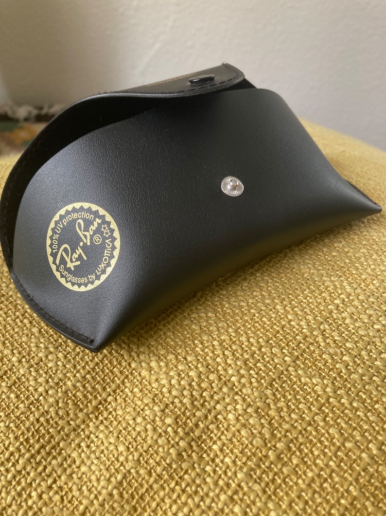 Op de afbeelding: Zwarte leren etui voor zonnebrillen met een gouden Ray-Ban logo met de tekst "100% UV protection Sunglasses by Luxottica"
