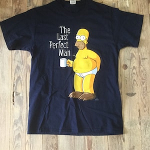 Könnte beinhalten: Ein marineblaues T-Shirt mit einem Cartoon-Bild von Homer Simpson, der eine Tasse hält. Der Text "The Last Perfect Man..." ist auf dem T-Shirt aufgedruckt.