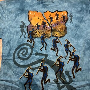 May include: Blue tie-dye t-shirt featuring a graphic of silhouetted figures playing instruments around a sun and spiral design. The figures are in dark blue, set against a tan and orange background. The shirt has a tag at the neckline.