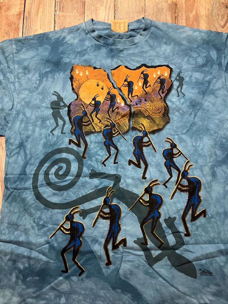 May include: Blue tie-dye t-shirt featuring a graphic of silhouetted figures playing instruments around a sun and spiral design. The figures are in dark blue, set against a tan and orange background. The shirt has a tag at the neckline.