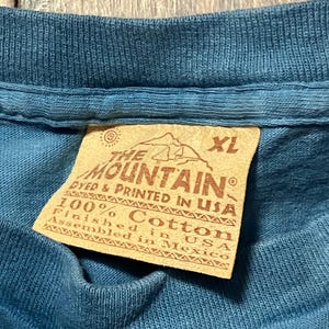 May include: Close-up of a blue XL t-shirt with a tag. The tag reads "THE MOUNTAIN" and "100% Cotton, Finished in USA, Assembled in Mexico."