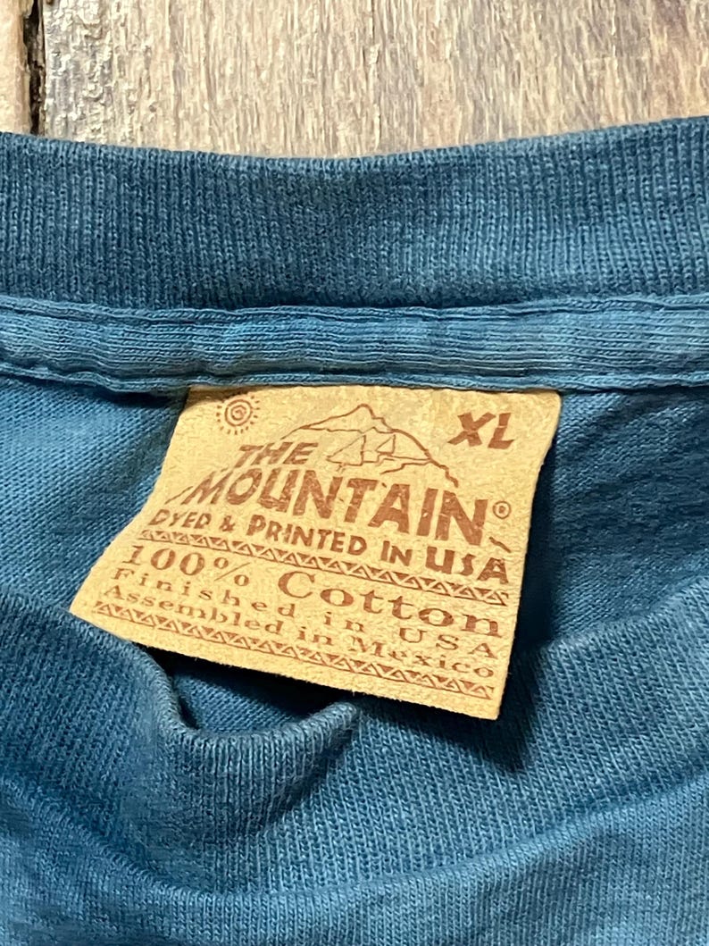 May include: Close-up of a blue XL t-shirt with a tag. The tag reads "THE MOUNTAIN" and "100% Cotton, Finished in USA, Assembled in Mexico."
