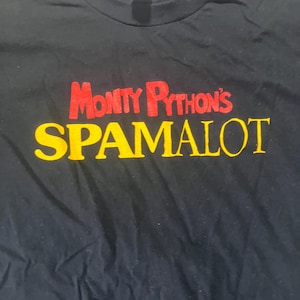 Może przedstawiać: Czarny t-shirt z napisem "Monty Python's SPAMALOT" w czerwonych i żółtych literach. Koszulka z okrągłym dekoltem wykonana jest z miękkiego materiału. Tekst jest wyśrodkowany na przodzie odzieży.