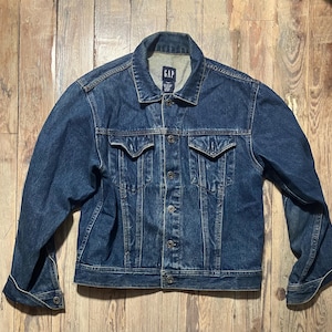 Puede incluir: Chaqueta vaquera azul con cierre de botones, dos bolsillos en el pecho y bolsillos laterales. La chaqueta tiene un cuello clásico y mangas largas. El nombre de la marca "GAP" es visible en el cuello interior.