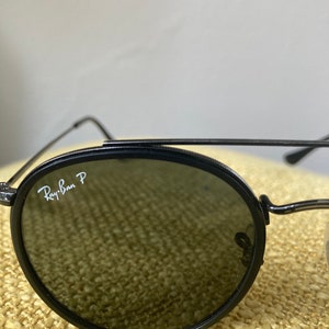 Op de afbeelding: Zwarte Ray-Ban P ronde zonnebril met donkere glazen. De zonnebril ligt op een beige, getextureerd oppervlak.