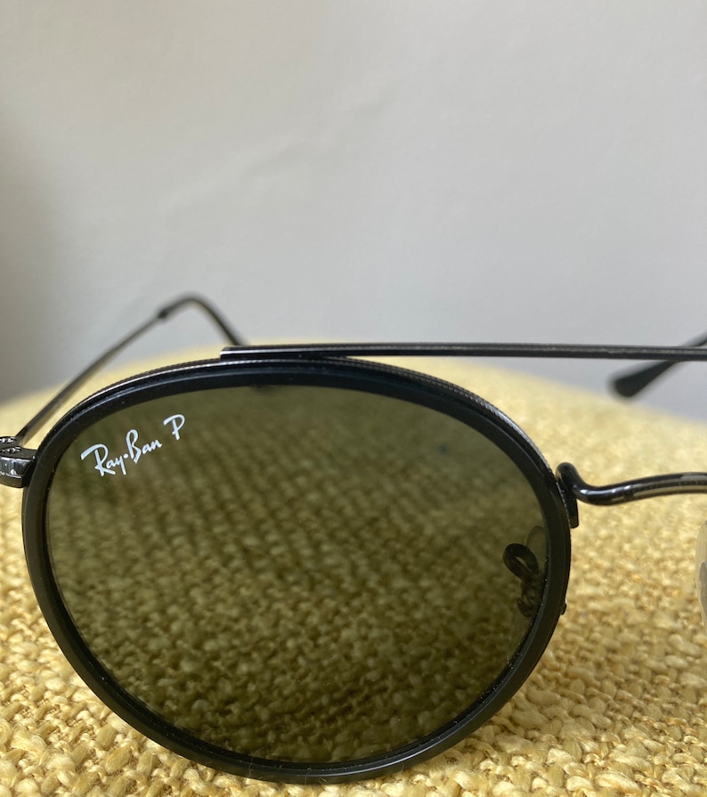 Op de afbeelding: Zwarte Ray-Ban P ronde zonnebril met donkere glazen. De zonnebril ligt op een beige, getextureerd oppervlak.