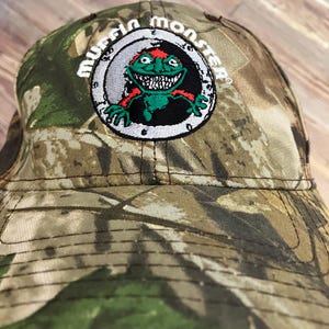 May include: Camouflage baseball cap with the text "Mussin Monster" embroidered on the front. The patch features a green monster with red accents. The cap has a curved brim and a classic six-panel design.