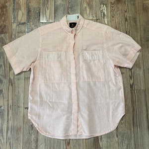 Puede incluir: Una camisa rosa claro de manga corta con rayas blancas verticales. Tiene dos bolsillos en el pecho y un cuello abotonado. La tela parece ligera, adecuada para climas cálidos.