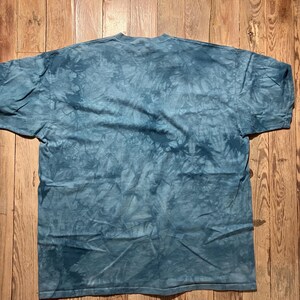 May include: A blue tie-dye t-shirt with a crew neck. The shirt is laid flat on a wooden surface. The fabric has a mottled pattern of light and dark blue hues, creating a textured appearance. The shirt's sleeves are short.