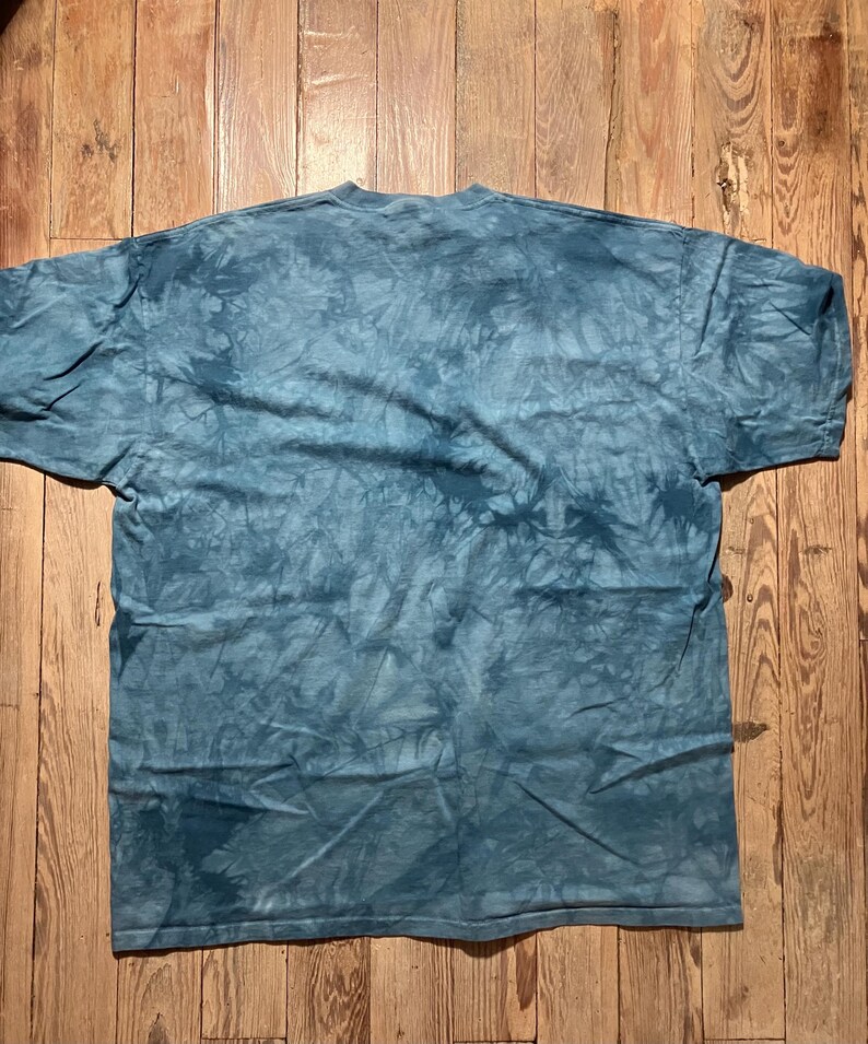 May include: A blue tie-dye t-shirt with a crew neck. The shirt is laid flat on a wooden surface. The fabric has a mottled pattern of light and dark blue hues, creating a textured appearance. The shirt's sleeves are short.